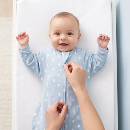 Bébé éveillé bras levés sur la table à langer, un parent ferme le zip de la gigoteuse TOG 1 bleue à gouttes