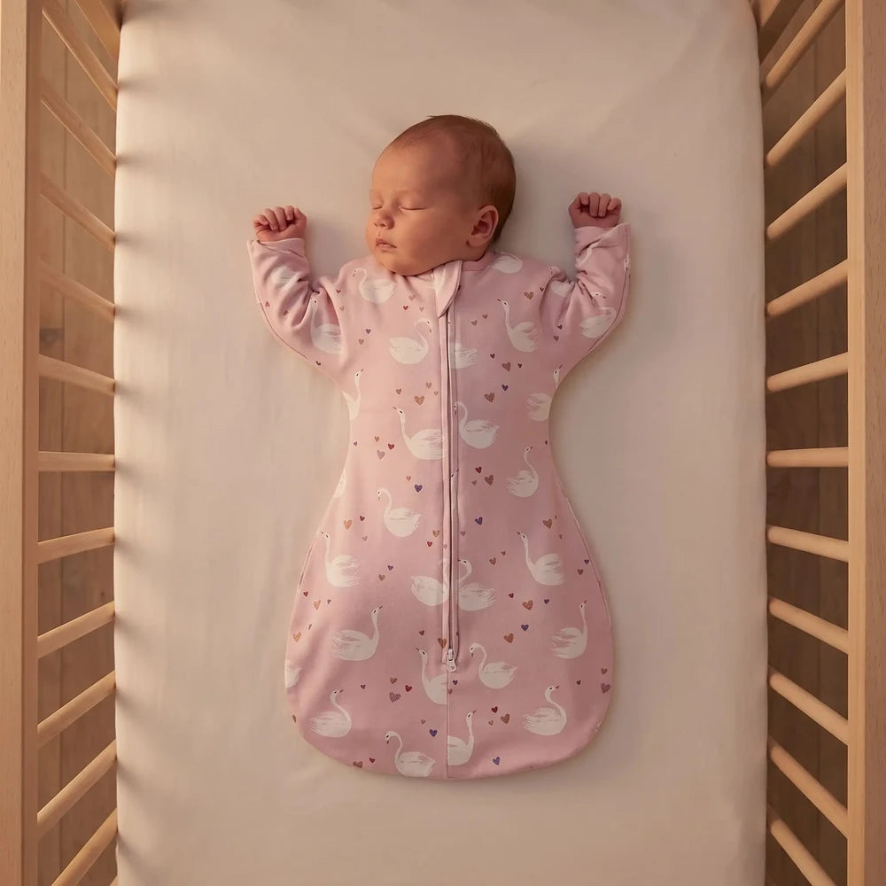 Nouveau-né endormi sur le dos, bras levés, en gigoteuse TOG 1 rose à cygnes dans un lit à barreaux vide